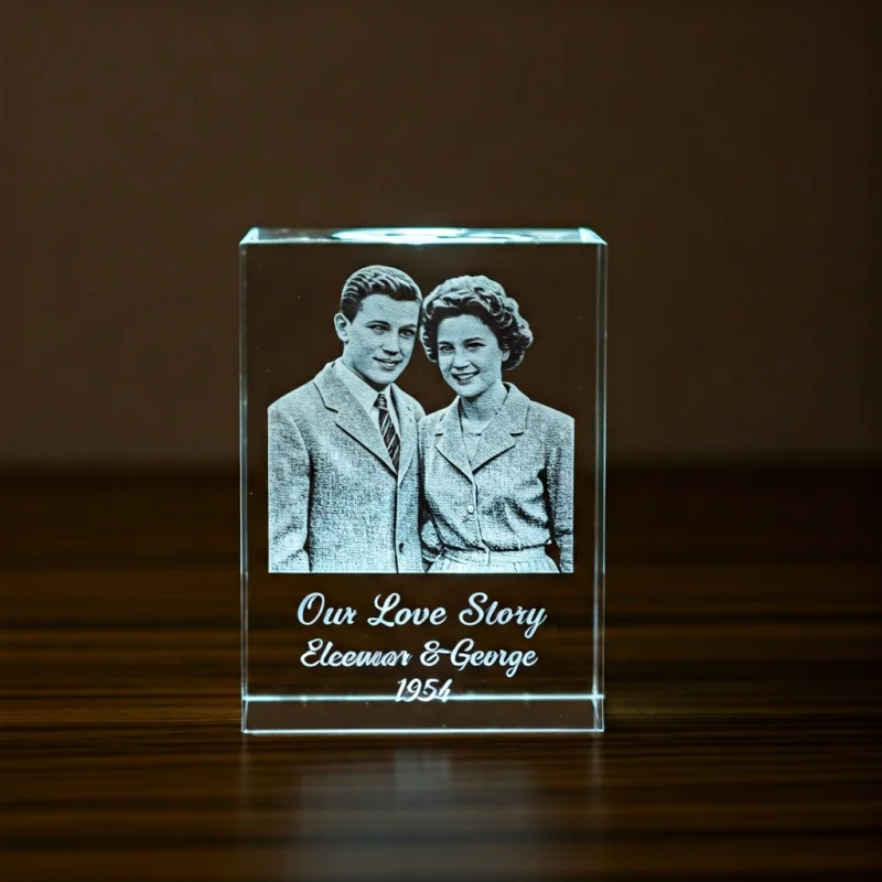 A personalized K9 crystal cube illuminated from below, showing a 2D laser-engraved image of a young couple from the 1950s, with custom text 'Our Love Story, Eleanor & George, 1954' at the base.